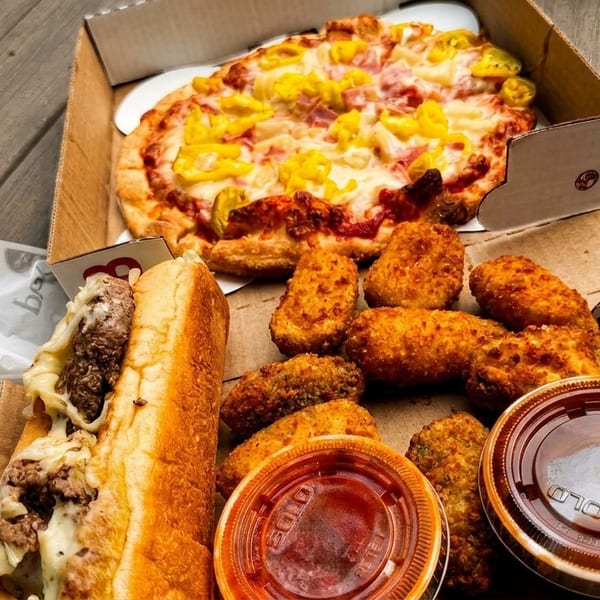 A Boston Pizza takeout box filled with a pizza, chicken wings, a Philly cheesesteak, and dipping sauces.